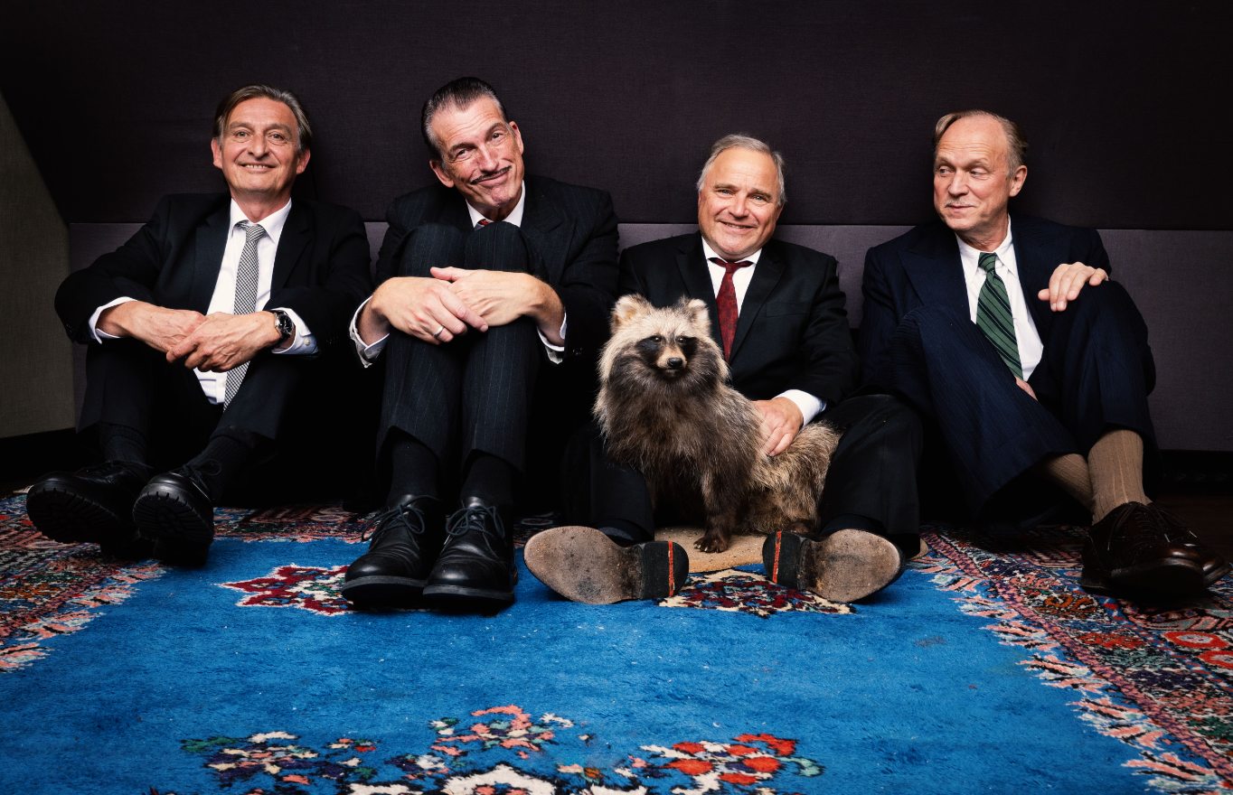 Ulrich Tukur und seine Band sitzen in eleganten Anzügen entspannt nebeneinander auf einem blauen Teppich und blicken lächelnd in die Kamera; in ihrer Mitte ein ausgestopfter Marderhund; die Szene wirkt humorvoll und ungezwungen.