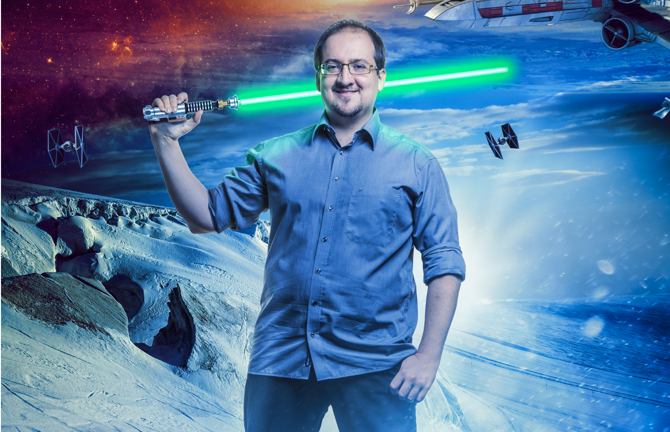 Der Wissenschaftler Sascha Vogel trägt ein kurzärmeliges, blaues Hemd und blickt in die Kamera. In seiner rechten Hand hält er ein Neonschwert, das hinter seinem Rücken grün erstrahlt. Im Hintergrund sieht man die Kraterlandschaft eines Planeten, buntes Licht im Universum und eine Weltraumsonde.