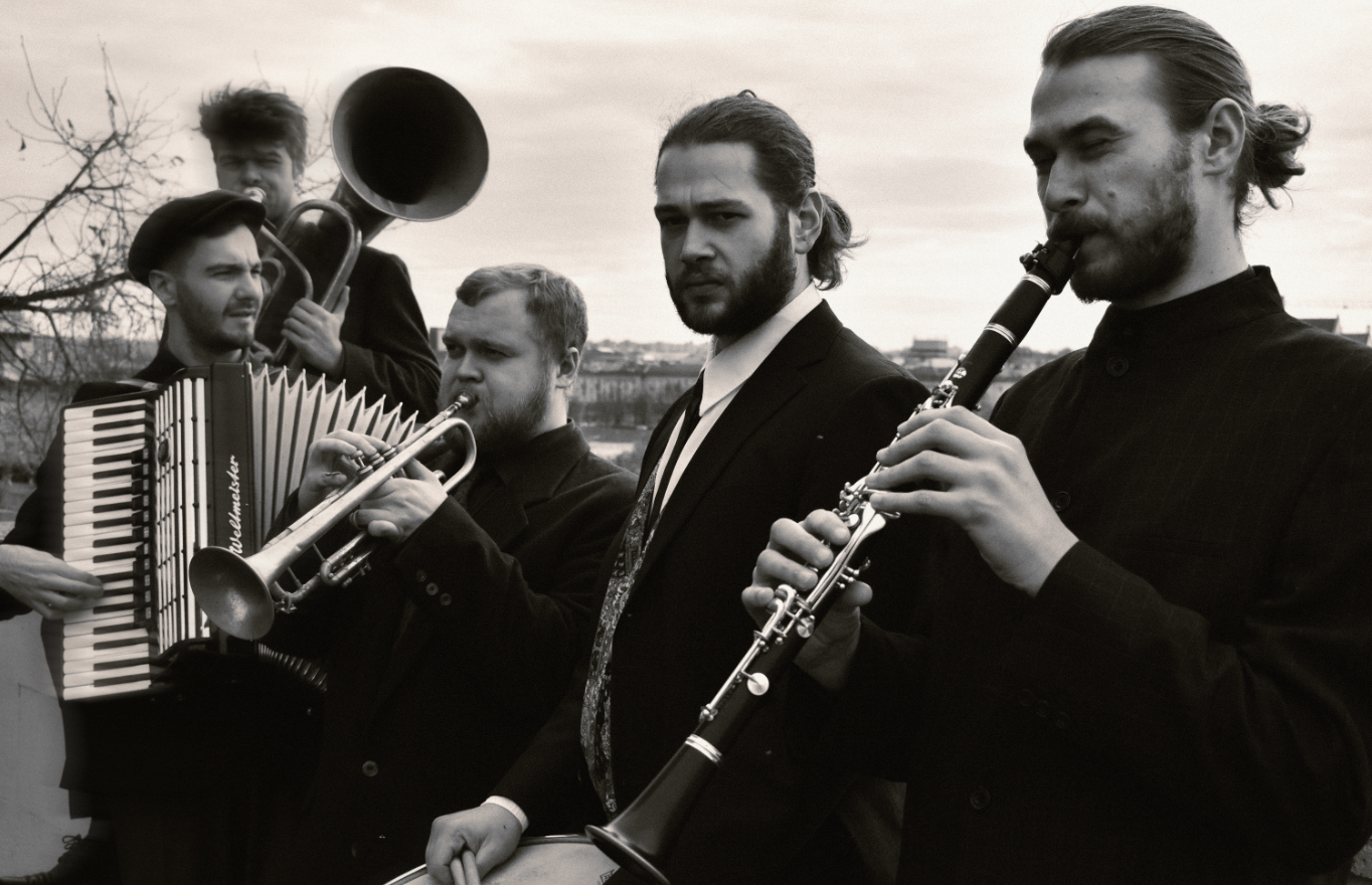 Schwarz-weiß Fotografie mit fünf Musikern in dunkler Kleidung mit ihren Instrumenten: Klarinette, Trommel, Trompete, Akkordeon und Tuba.