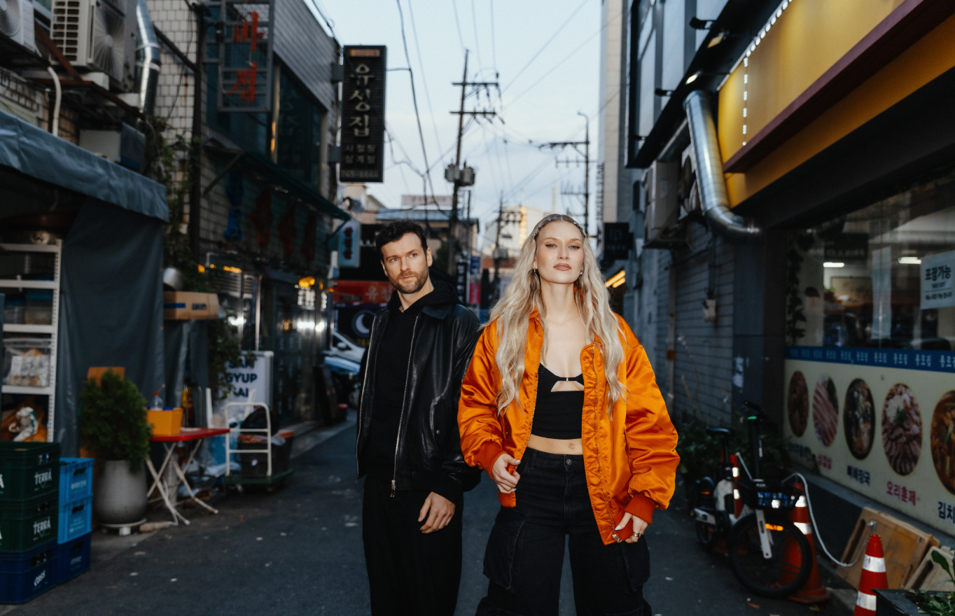 Die Musiker der Band stehen in einer schmalen Straße mit Geschäften, Schildern und Lichtern. Carolin rechts im Bild trägt eine leuchtend orangene Bomberjacke, Daniel links von ihr ist ganz in schwarz gekleidet.